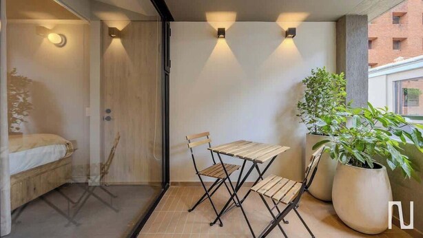 A balcony with a wooden table and chairs, a potted plant, and wall-mounted lights. - NOK Designer Apartment 1BR in Zona Rosa (Bogotá)