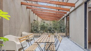Een overdekt terras met een houten tafel en stoelen, een witte bank en een groene plant.