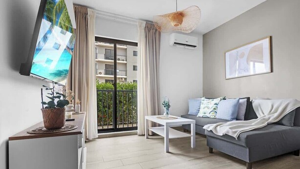 A modern living room with a grey sofa, a white coffee table, and a flat-screen TV on a wooden stand. - NOK Amazing 1BR Apartment Jardines 3, Punta Cana (Punta Cana)