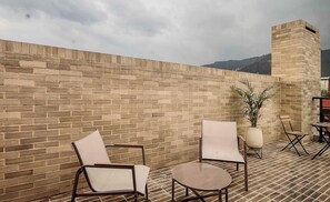Eine Dachterrasse mit Backsteinmauern, zwei Stühlen, einem kleinen Tisch, einem Blumentopf und einem Klappsessel.