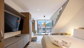 Ein modernes Hotelzimmer mit Fernseher, einem Bett mit Handtüchern, einem Schreibtisch und Blick auf Gebäude durch große Fenster.