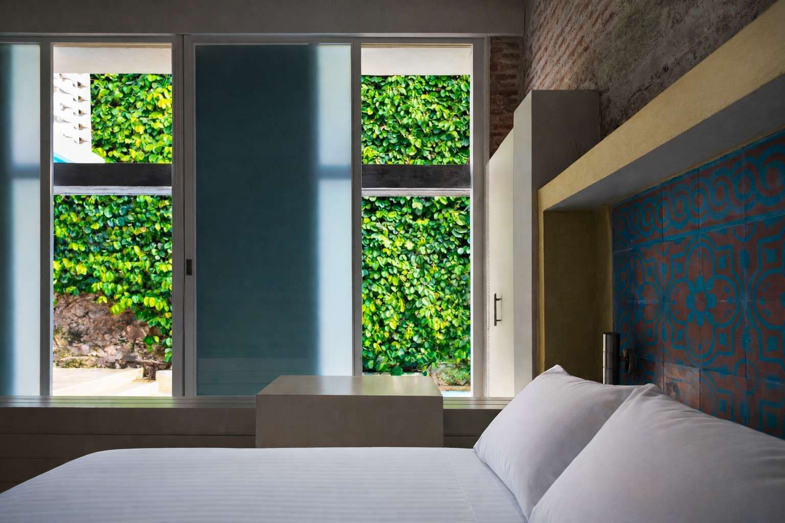 A bedroom with a bed, a window with a view of greenery, and a patterned wall.