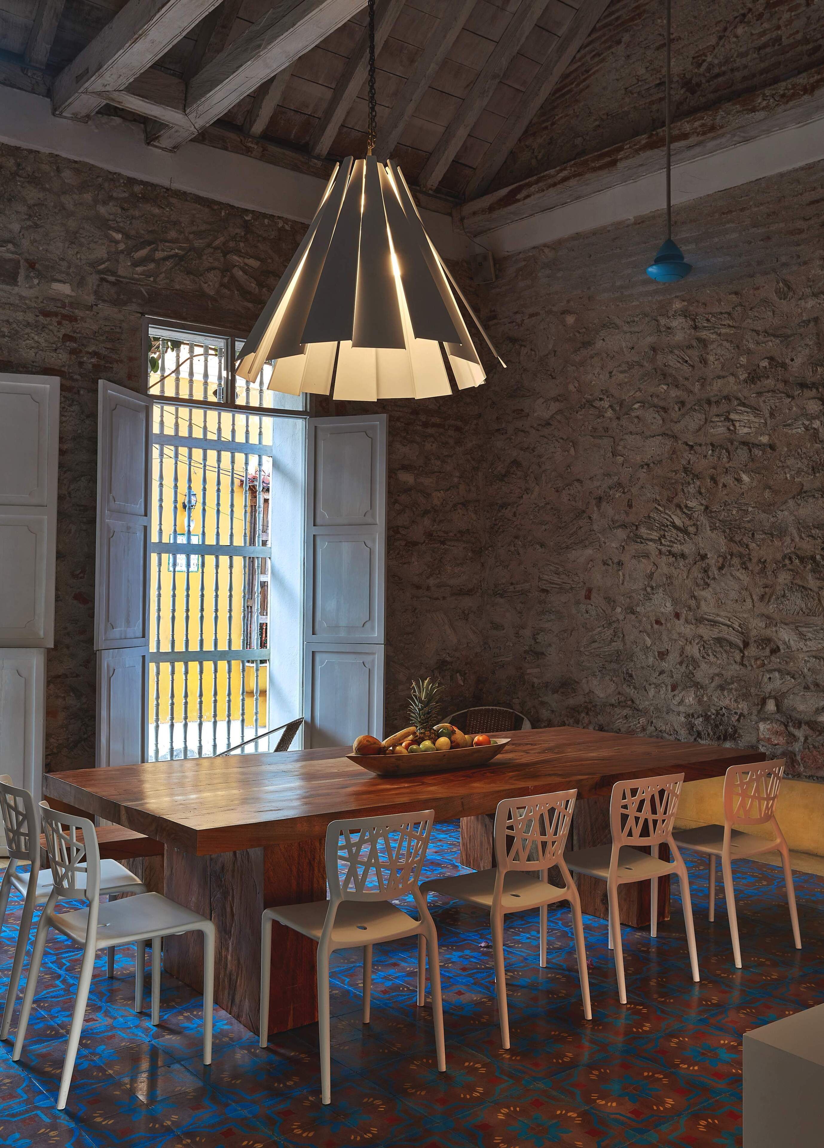 A rustic dining area with a wooden table, white chairs, a hanging pendant light, and a view through a barred window.