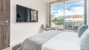 A modern bedroom with a large bed, a flat-screen TV mounted on the wall, a chair, and a large window offering a view of buildings and greenery. - NOK Designer 2BR in Usaquén (Bogotá)