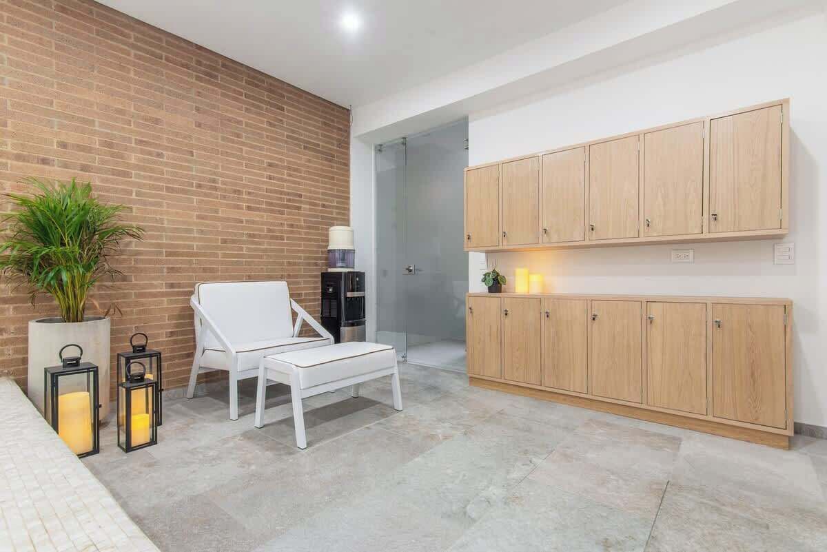 A modern living room with a white sofa, a small table, and a potted plant. There are wooden cabinets and a glass door leading to another room.