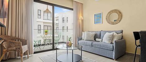 A modern living room with a grey sofa, a wicker chair, a glass coffee table, and a view of a building through large windows.