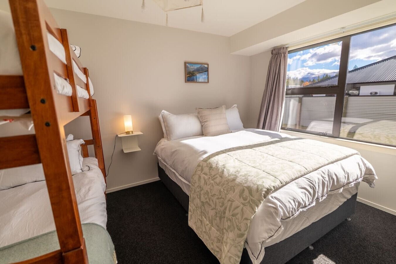 A bedroom with a bunk bed, a single bed, a bedside lamp, a window with a view, and a painting on the wall.