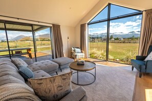 A modern living room with a large sofa, a coffee table, and a view of mountains through large windows. - Ben Ohau Vista Lodge | Stunning Mountain Views (Twizel)