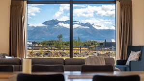 Ein Zimmer mit großem Fenster und Blick auf die Berge, einem Essbereich mit Tischen und Stühlen sowie einem blauen Sofa.