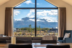 A room with a large window offering a mountain view, a dining area with tables and chairs, and a blue sofa. - Ben Ohau Vista Lodge | Stunning Mountain Views (Twizel)