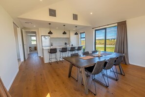 A modern kitchen with bar stools, a dining table with chairs, and a large window with a view. - Ben Ohau Vista Lodge | Stunning Mountain Views (Twizel)