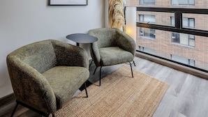 Een modern interieur met twee groene fauteuils, een kleine ronde tafel, een inlijsting foto aan de muur en uitzicht op een bakstenen gebouw door grote ramen.