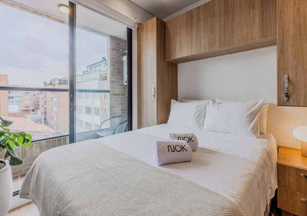 A hotel room with a large bed, a wooden headboard, a bedside table with a lamp, and a view of buildings through a large window. - NOK Stylish 1BR Apartment in North Bogota (Bogotá)