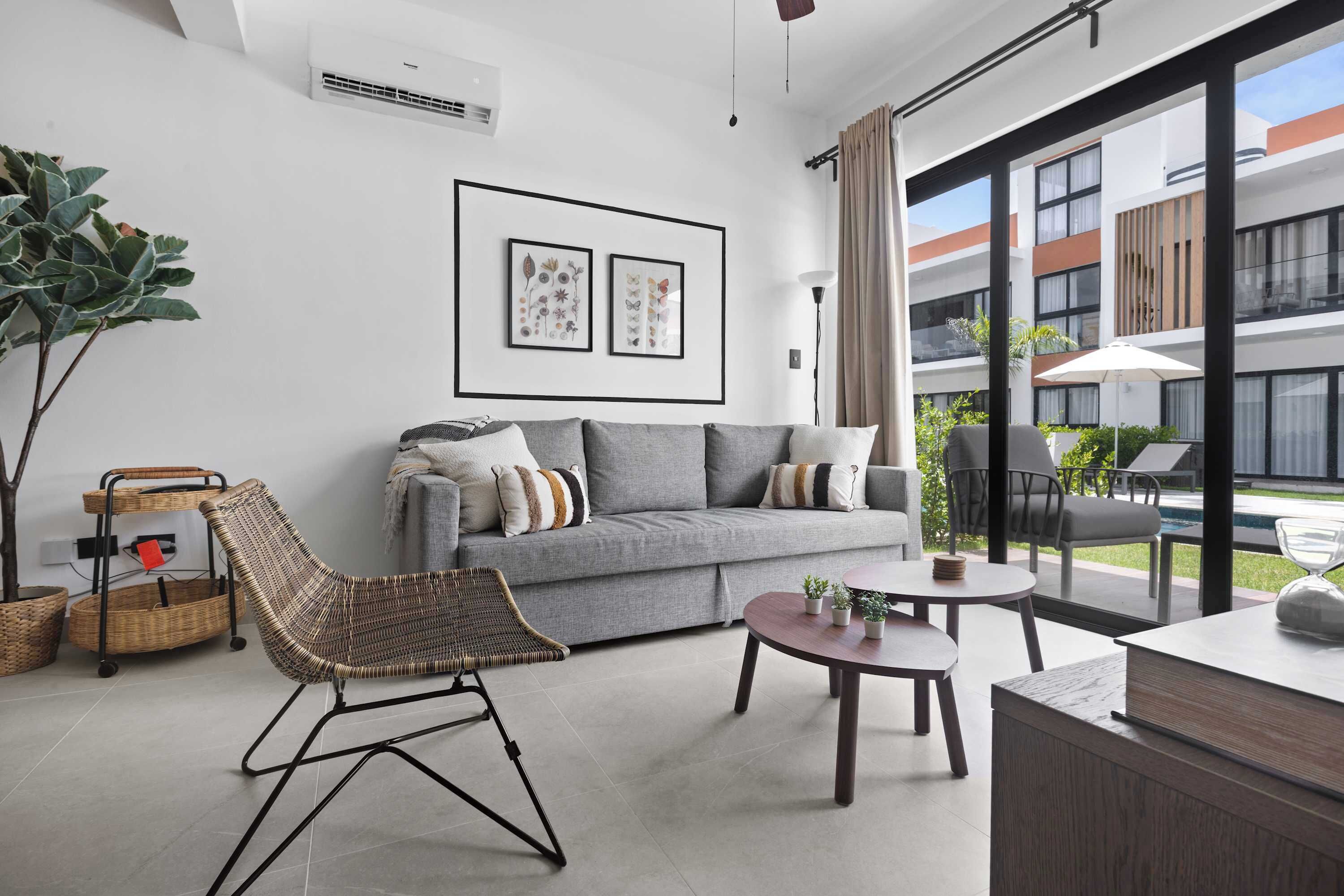 Un salon moderne avec un canapé gris, un fauteuil en osier et une table basse ronde. Les murs sont ornés de tableaux encadrés, il y a une grande fenêtre avec des rideaux et un balcon aménagé avec du mobilier d’extérieur.