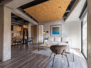 Un salon moderne avec un plafond en bois, un canapé blanc, une table basse et un coin repas avec des chaises, et une cuisine en arrière-plan.