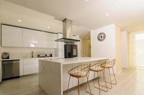 A modern kitchen with white cabinetry, a stainless steel dishwasher, a central island with bar stools, and a clock on the wall. - NOK Luxury 4BR in Usaquén (Bogotá)