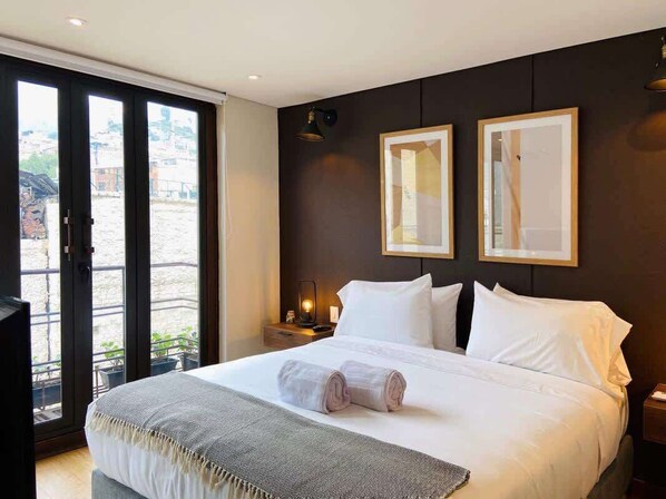 A hotel room with a large bed, two bedside tables, a nightstand, and a sliding glass door leading to an outdoor area. - NOK Great Studio (Bogotá)