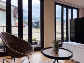A room with a large window, a wicker chair, a small table with a plant, and a TV on the right. - NOK Great Studio (Bogotá)