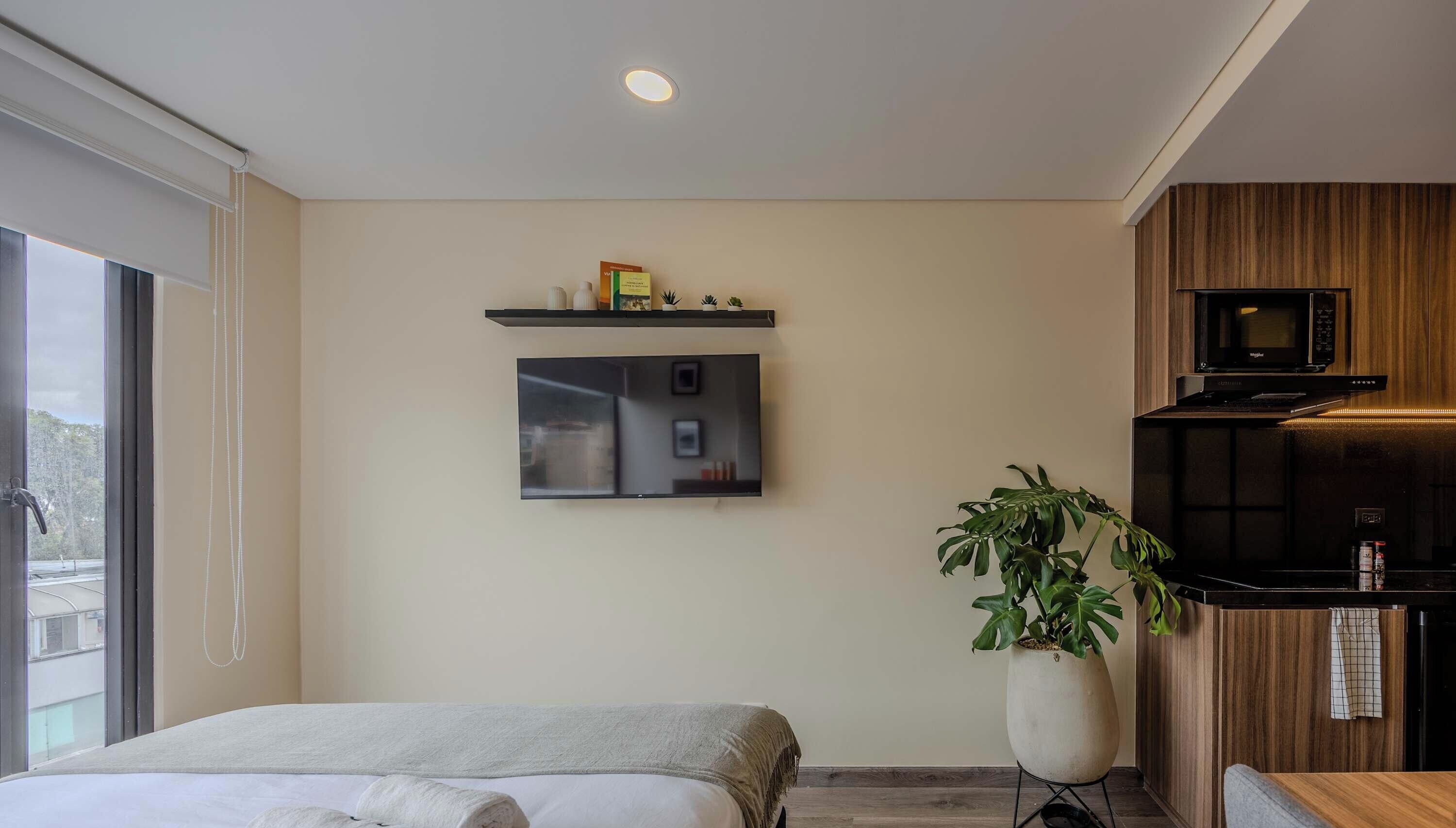Ein modernes Hotelzimmer mit Bett, einem Wand-Fernseher, einer kleinen Küchenzeile mit Mikrowelle und einem großen Fenster.