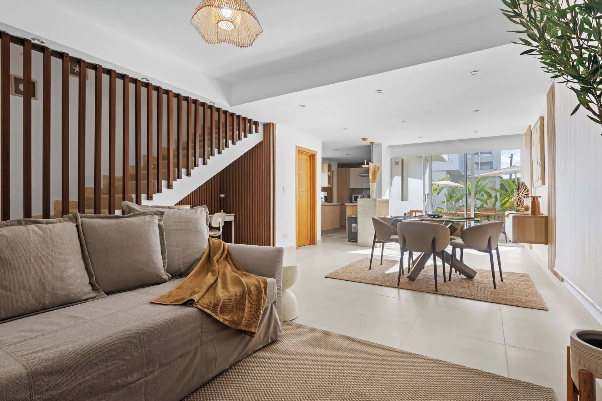 A modern living room with a sofa, a dining table with chairs, a staircase, and a kitchen area.
