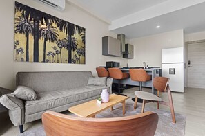 A modern living room with a grey sofa, wooden coffee table, and orange chairs. A kitchen area is visible in the background with a microwave, stove, and cabinets. - NOK Cozy 1BR Apartment in Panorama Lake-punta Cana (Punta Cana)