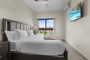 A bedroom with a bed, a nightstand, a lamp, a ceiling fan, an air conditioning unit, a TV, and a window with a view of buildings and greenery. - NOK Cozy 1BR Apartment in Panorama Lake-punta Cana (Punta Cana)