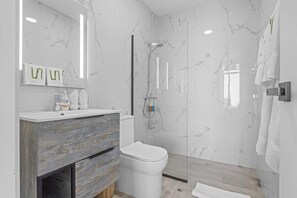 A modern bathroom with a marble wall, a wooden vanity, a white toilet, and a glass shower enclosure. - NOK Cozy 1BR Apartment the Towers By Vistacana (Punta Cana)