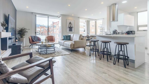 Un appartement moderne comprenant un espace de vie, une cuisine et un coin repas. Il est doté d’une grande fenêtre, d’un canapé, d’une table basse et d’un bar avec des tabourets.