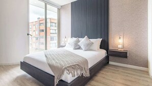 Une chambre moderne avec un grand lit, une table de chevet avec une lampe et une vue sur les immeubles par une grande fenêtre.