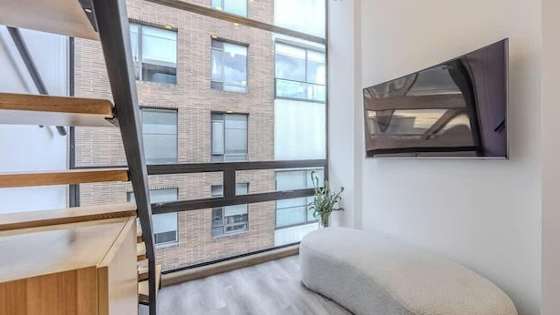 A modern apartment with a wooden staircase, a large window with a view of a brick building, a white sofa, and a mounted TV. - NOK Luxury Loft in Chico Norte (Bogotá)