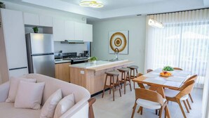 A modern kitchen with white cabinets, a central island, and a dining area with wooden chairs and a table. - NOK Classy 3BR Villa by Vistacana Rentals (Punta Cana)
