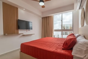 Um quarto com cama, televisão na parede, ventilador de teto e uma janela grande.