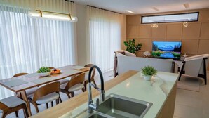 A modern kitchen with a central island, a dining area with wooden chairs, a built-in TV, and a large window with blinds. - NOK Classy 3BR Villa by Vistacana Rentals (Punta Cana)
