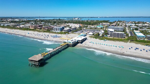 An aerial view of a beach with a pier extending into the water, surrounded by buildings and beach umbrellas. - Beachside Ground Floor Walkout. Heated Pool. BBQ Grills ☀️ (Cocoa Beach)