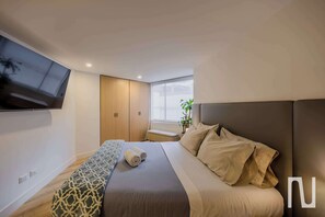 A modern hotel room with a bed, a bench, a TV mounted on the wall, and a window with a view. - NOK Beautiful 3BR Apt in Zona Rosa (Bogotá)