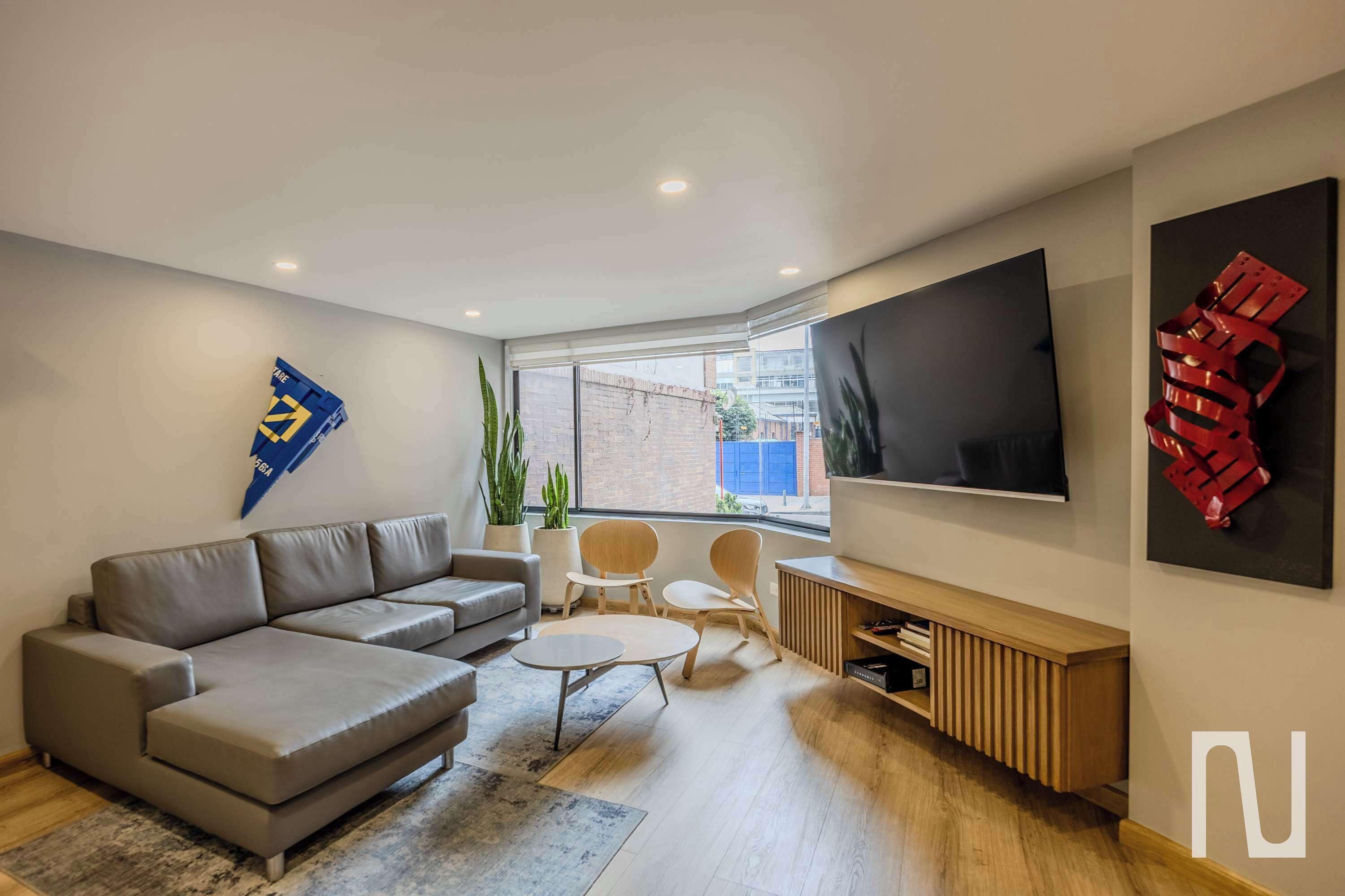 A modern living room with a grey sofa, a coffee table, and a TV mounted on the wall. There are two chairs and a small table.