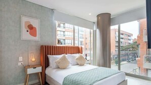 A modern bedroom with a large bed, a bedside table with a lamp, a framed abstract artwork on the wall, and a view of buildings through large windows. - NOK Amazing 1BR in Usaquén (Bogotá)