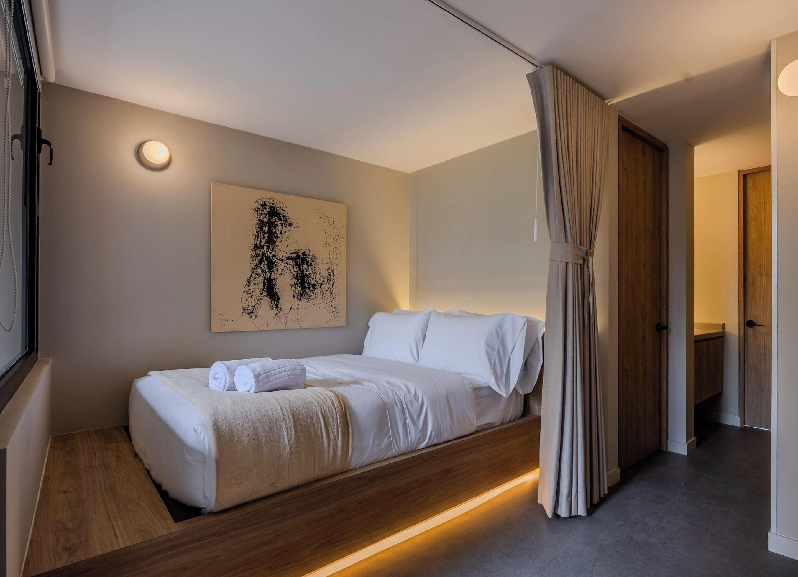 Quarto de hotel moderno com uma cama, uma obra de arte na parede, uma mesa de cabeceira e uma cortina.