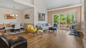 A spacious living room with a dining area, a sofa, a yellow armchair, and a balcony with a view of trees. - Modern Lakeview Escape: 2BR Golfers’ Retreat (Raleigh)