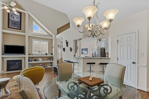 A modern living room with a glass dining table, comfortable seating, a fireplace, and a kitchen area in the background.