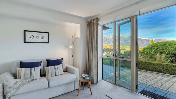 Ein modernes Wohnzimmer mit einer Couch, einem kleinen Tisch, einer Stehlampe und einer großen Glasschiebetür, die auf einen Balkon mit Bergblick führt.