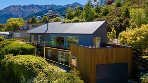 Ein modernes Haus mit großem Glasbalkon, umgeben von üppiger Vegetation und Bergen im Hintergrund.