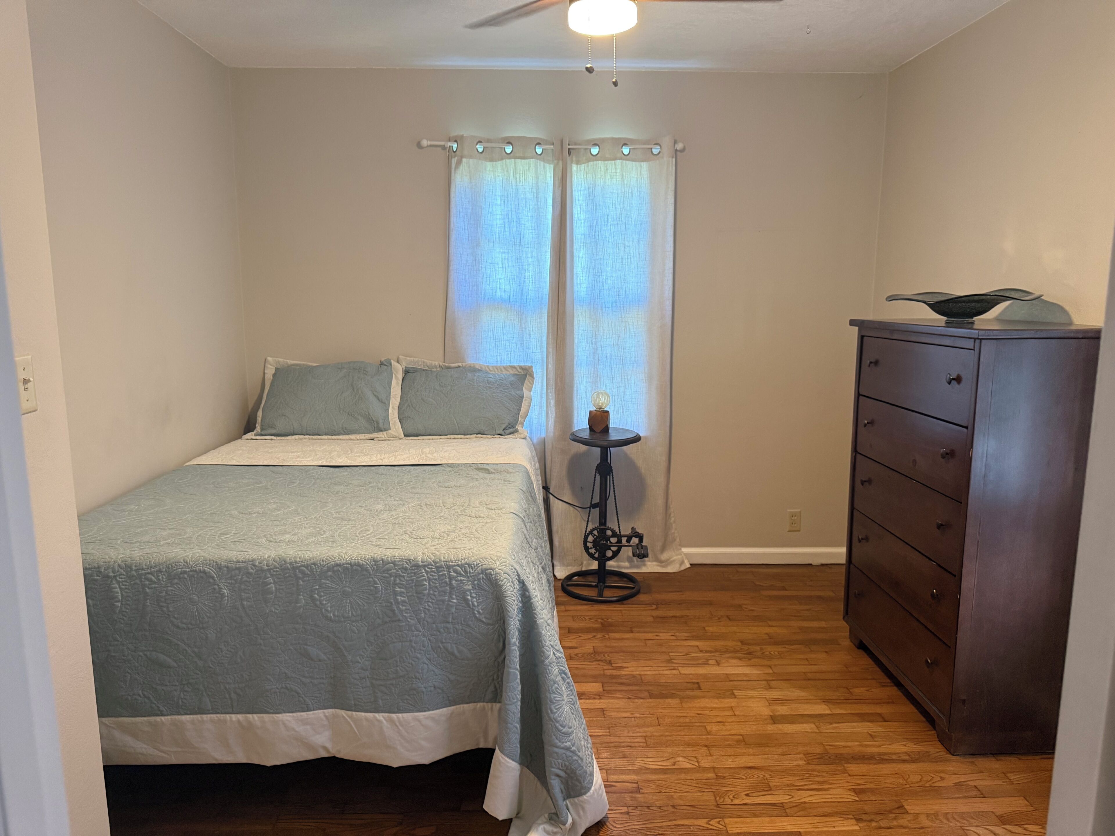Une chambre à coucher avec un lit, une commode, une petite table avec une lampe et une fenêtre avec des rideaux.