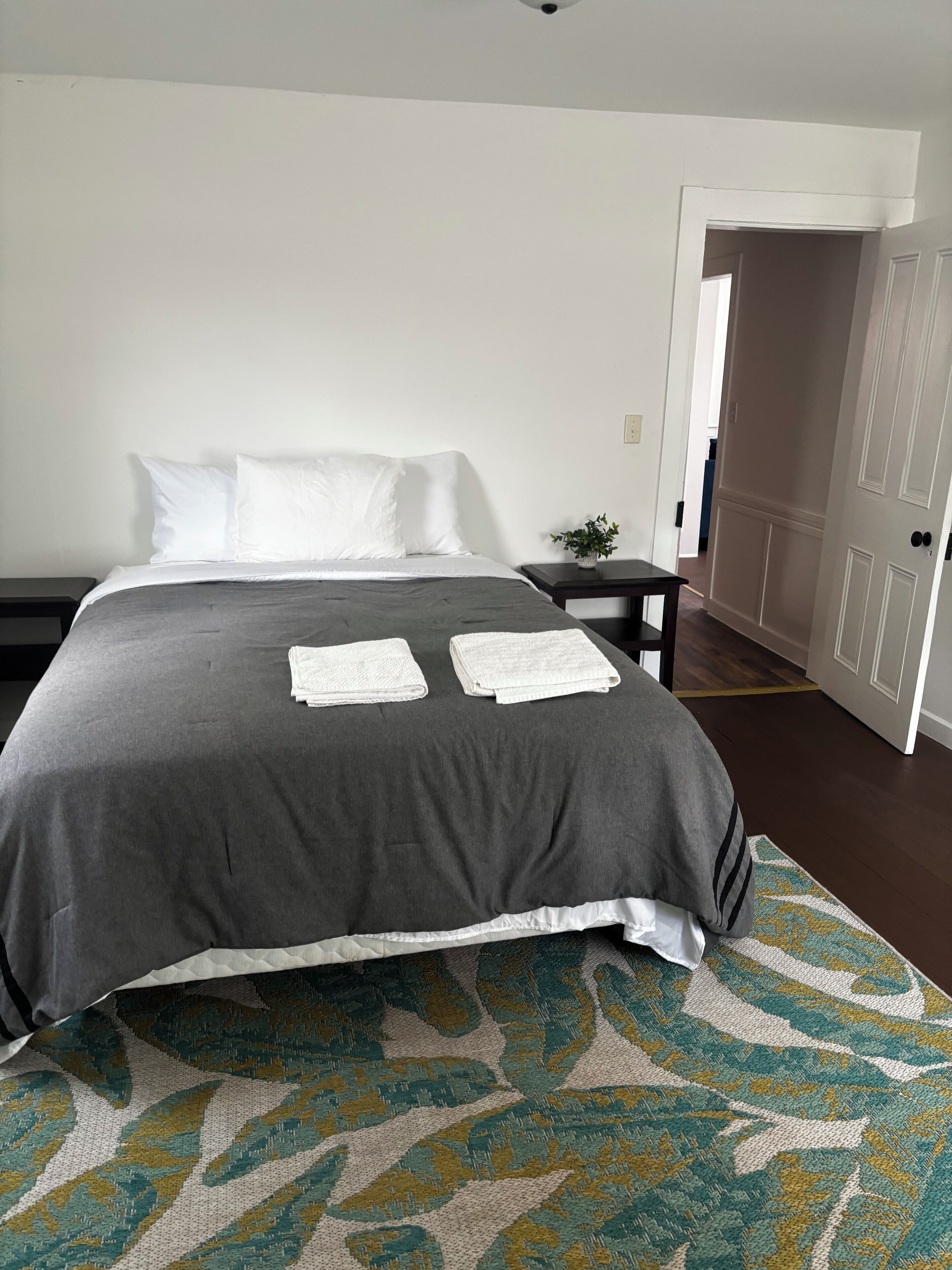 A neatly made bed with a grey bedspread and white pillows, a small bedside table with a plant, and a patterned rug on the floor.