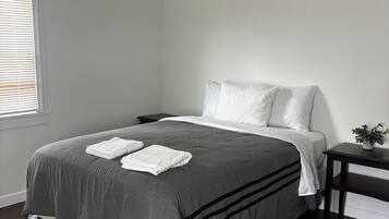 Una cama bien tendida con una colcha gris y almohadas blancas, una mesita de noche con una planta y una alfombra estampada sobre un suelo oscuro.