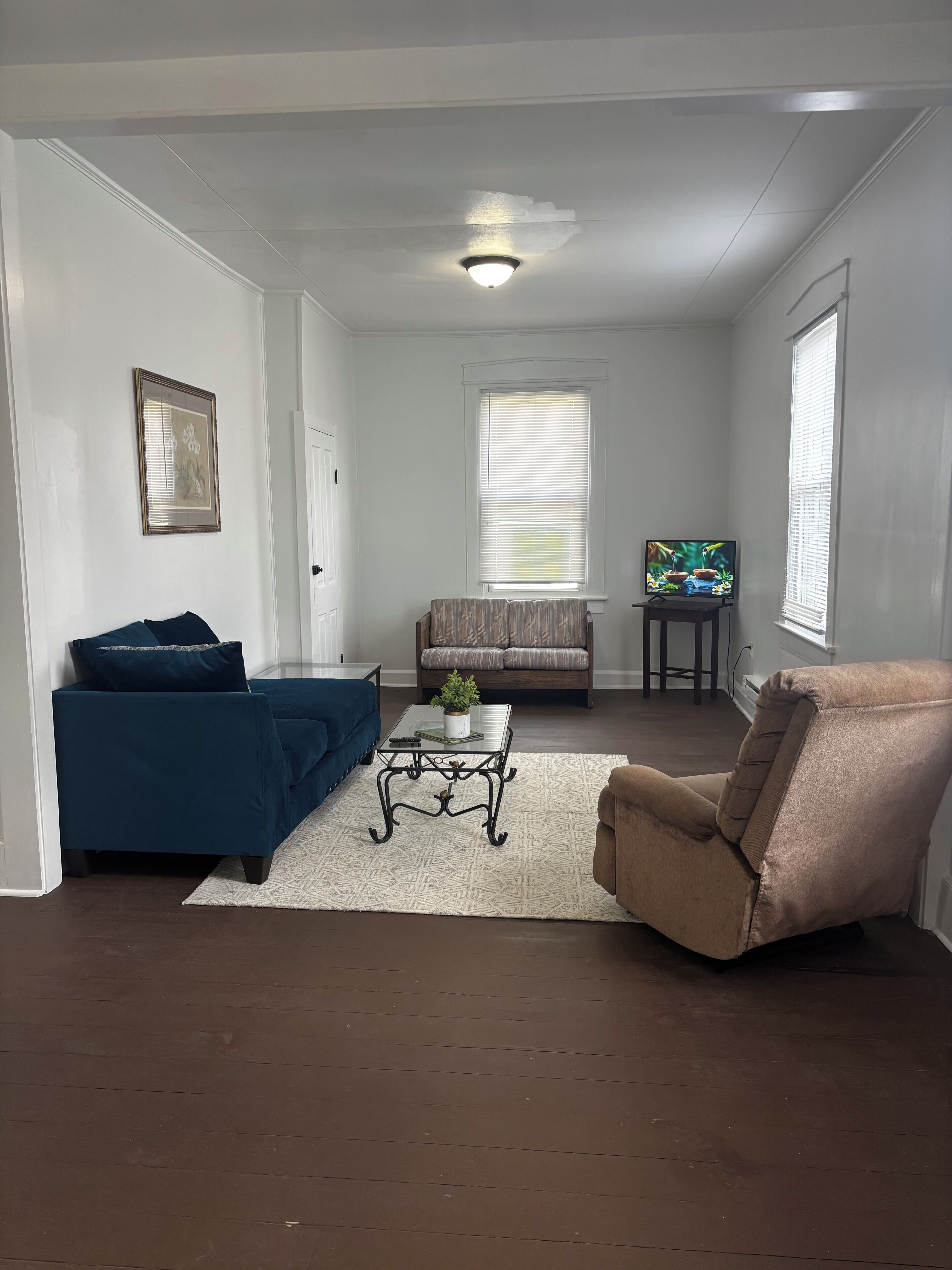 Una sala de estar con un sofá azul, un sillón beige, una mesa de centro de vidrio con una planta, un televisor sobre un mueble bajo y una ventana con persianas.