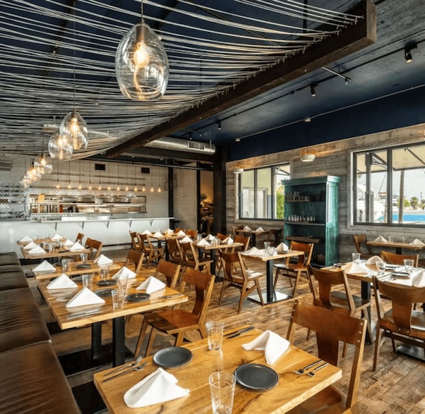 Un restaurante con mesas y sillas de madera, lámparas colgantes y vistas amplias a través de grandes ventanales.
