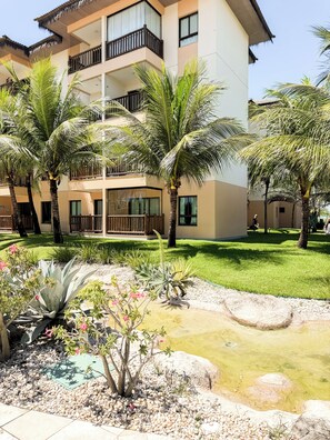 Um hotel com vários andares, varandas, palmeiras e um jardim paisagístico com pedras e plantas.