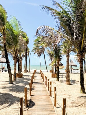 A wooden pathway leading to a beach with palm trees and a thatched-roof structure. - Resort Vg Sun Cumbuco, Apartment 112 (Caucaia)