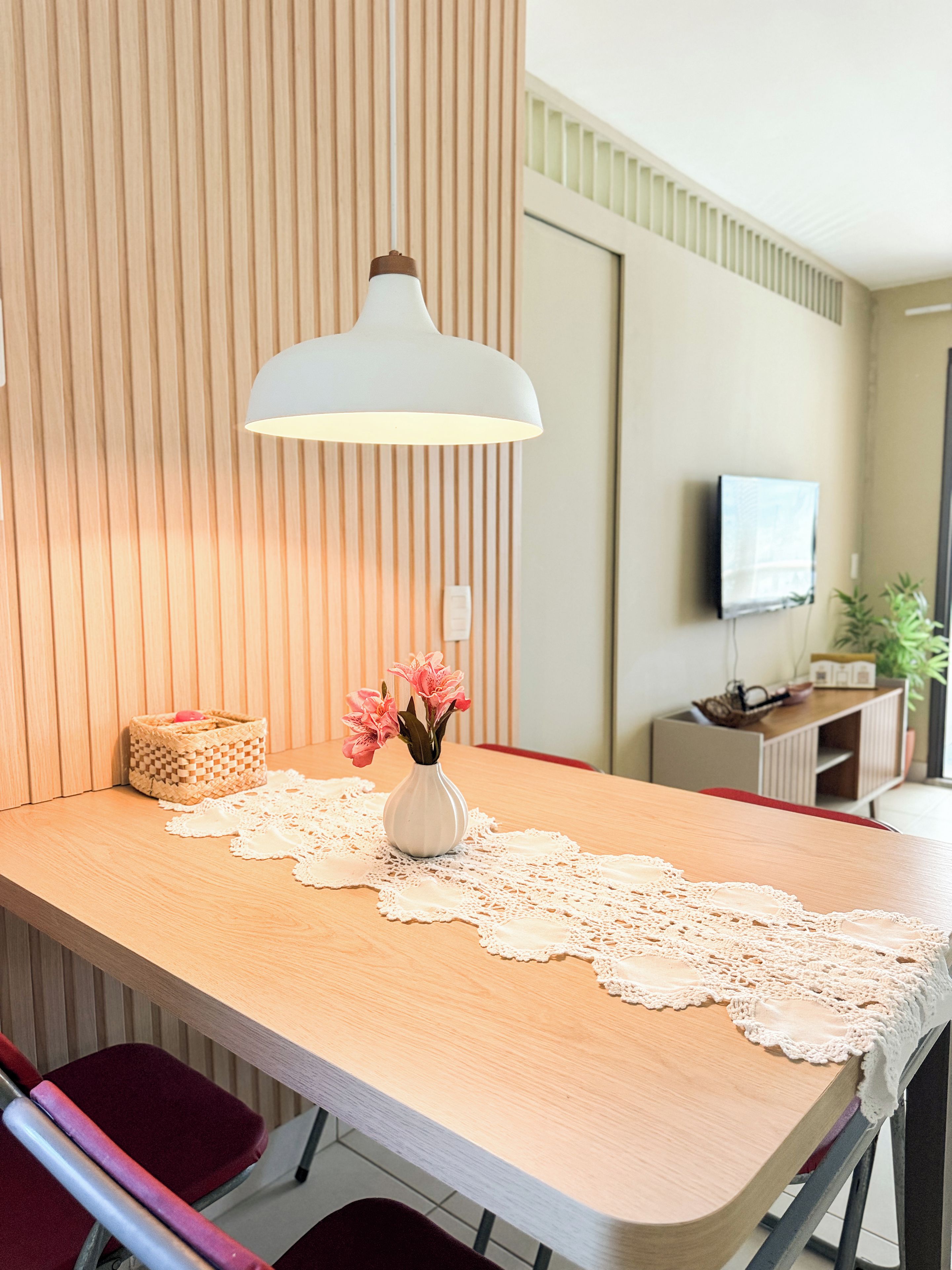 A dining area with a wooden table, a white vase with flowers, a lace table runner, and a hanging pendant light.